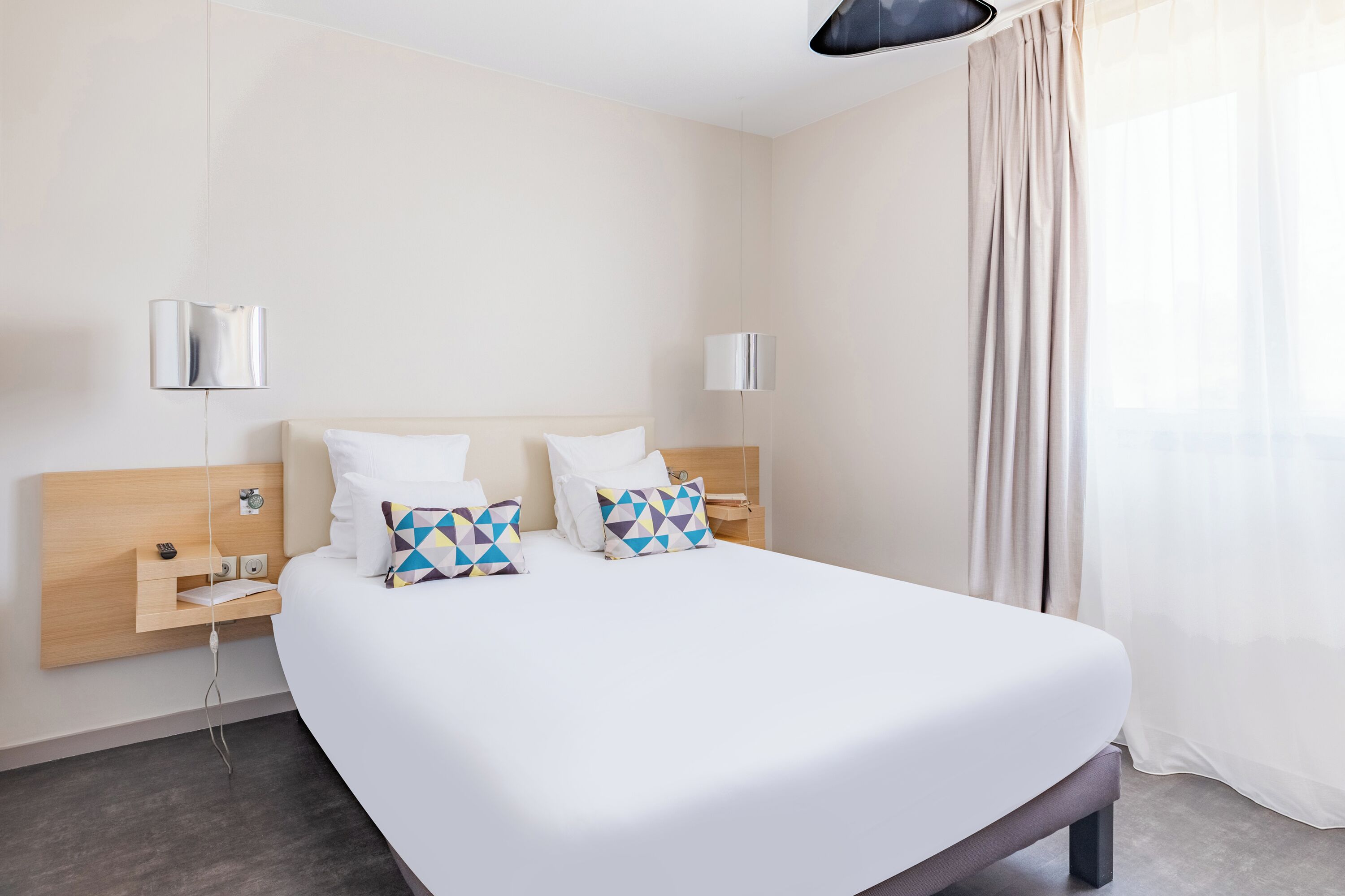 A modern hotel room with a large bed, two bedside tables, a wooden headboard, and a large window with curtains.