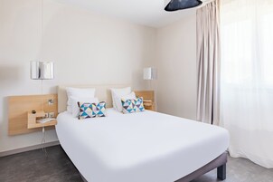 A modern hotel room with a large bed, two bedside tables, a wooden headboard, and a large window with curtains.