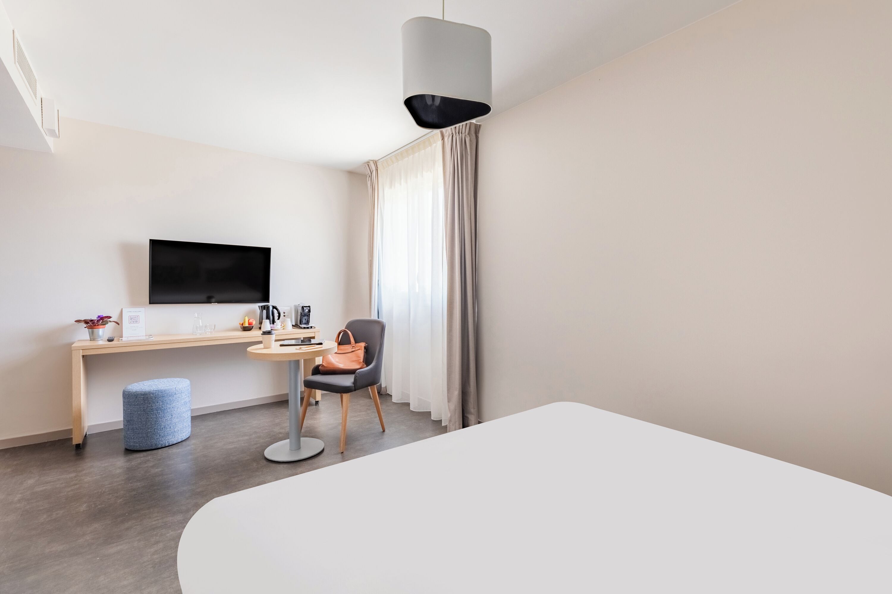A modern hotel room with a flat-screen TV, a desk with a chair, a round table, and a large bed.