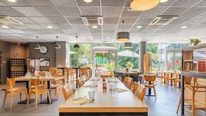 A spacious restaurant with long wooden tables and chairs, a modern ceiling design, and large windows offering a view of the outdoors. - Appart'City Confort Lyon Cité Internationale (Caluire-et-Cuire)
