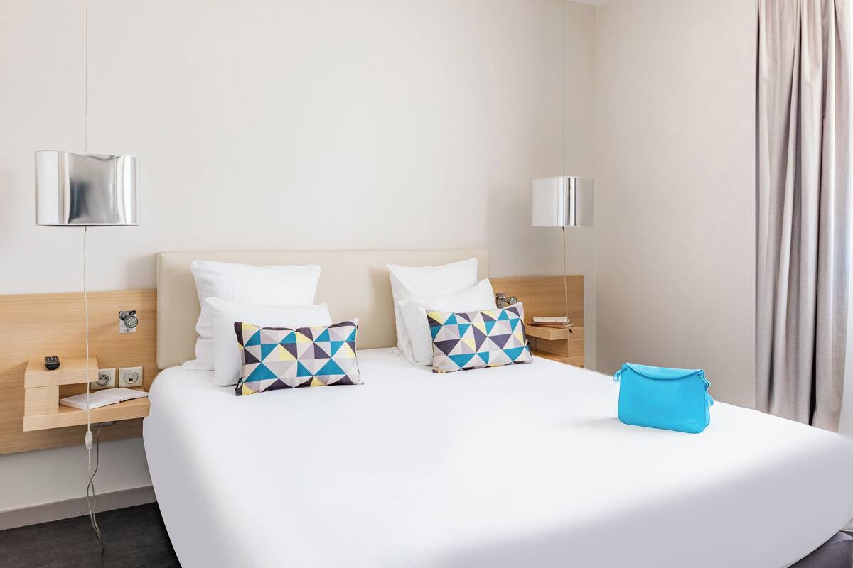 A hotel room with a large bed, two pillows, a bedside table, a lamp, and a blue bag on the bed.