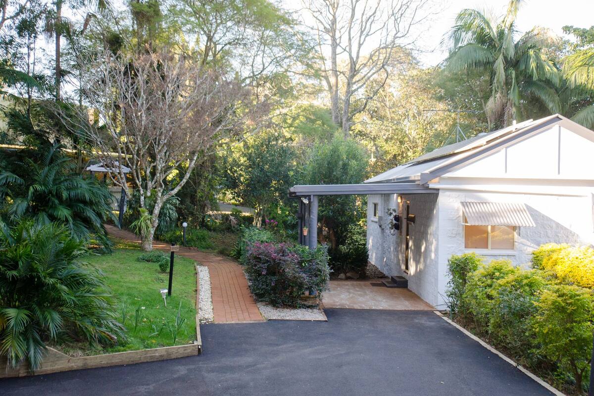 A house with a covered porch, a paved driveway, and a garden with various plants and trees.