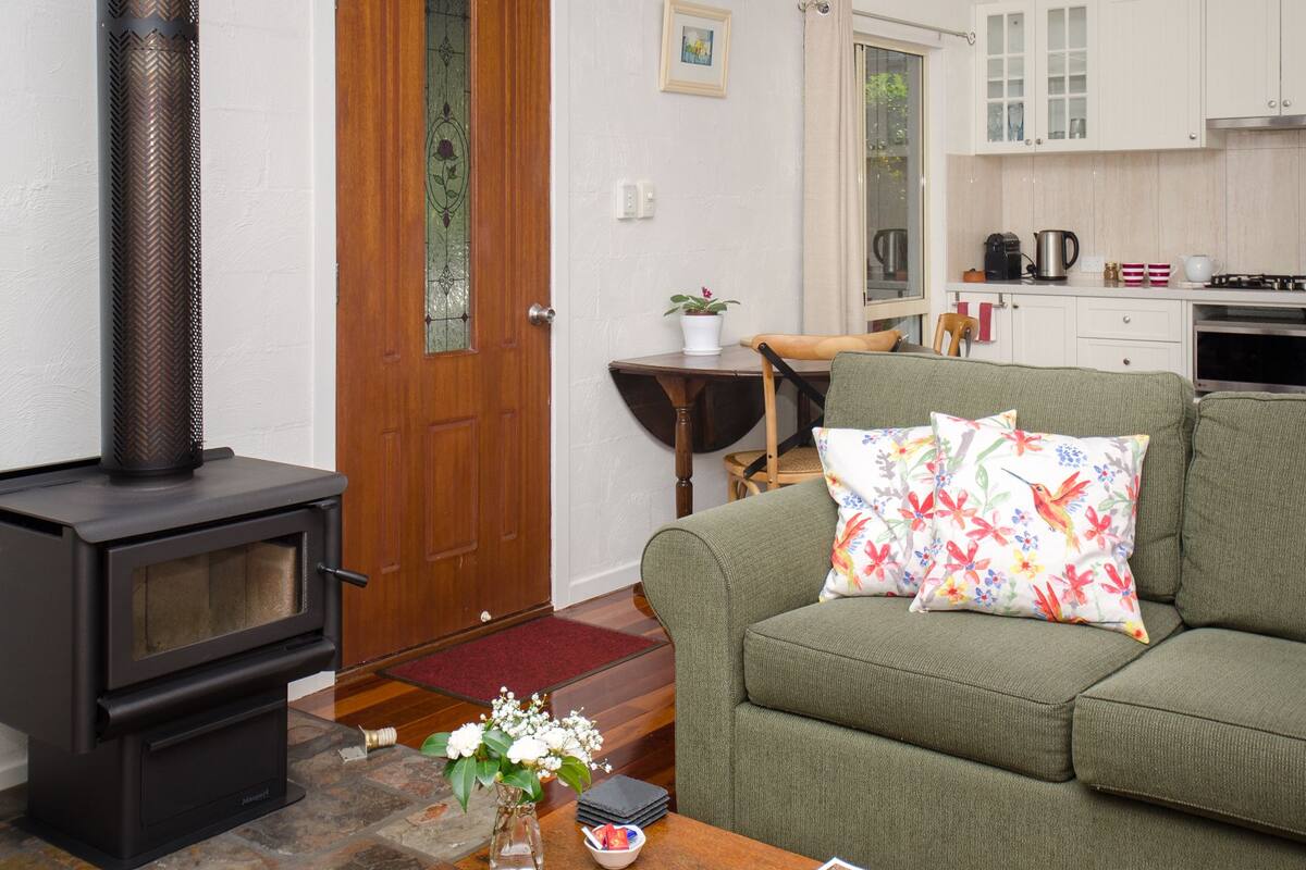 A living room with a fireplace, a sofa with floral cushions, a dining table, and a kitchen in the background.