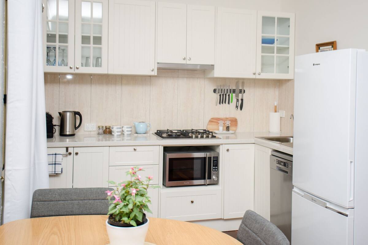 A modern kitchen with white cabinetry, a gas stove, an oven, a microwave, and a dishwasher. There is a dining table with chairs and a potted plant.