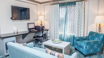 A hotel room with a flat-screen TV, a desk with a computer, a blue armchair, a sofa, and a dining table with a bowl.