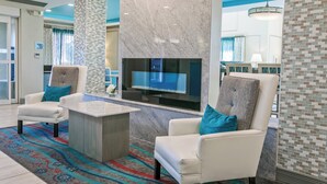Eine moderne Lobby mit Kamin aus Marmor, zwei weißen Ledersesseln mit türkisblauen Kissen, einem Holztisch und einem farbenfrohen Teppich.