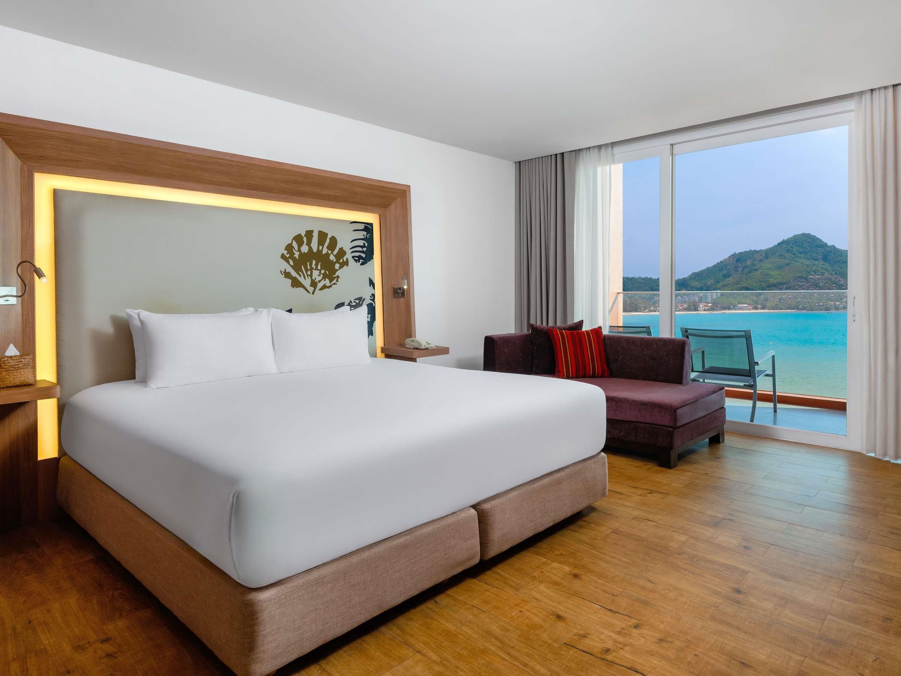 A hotel room with a large bed, a sofa, a nightstand, a phone, and a view of the sea.