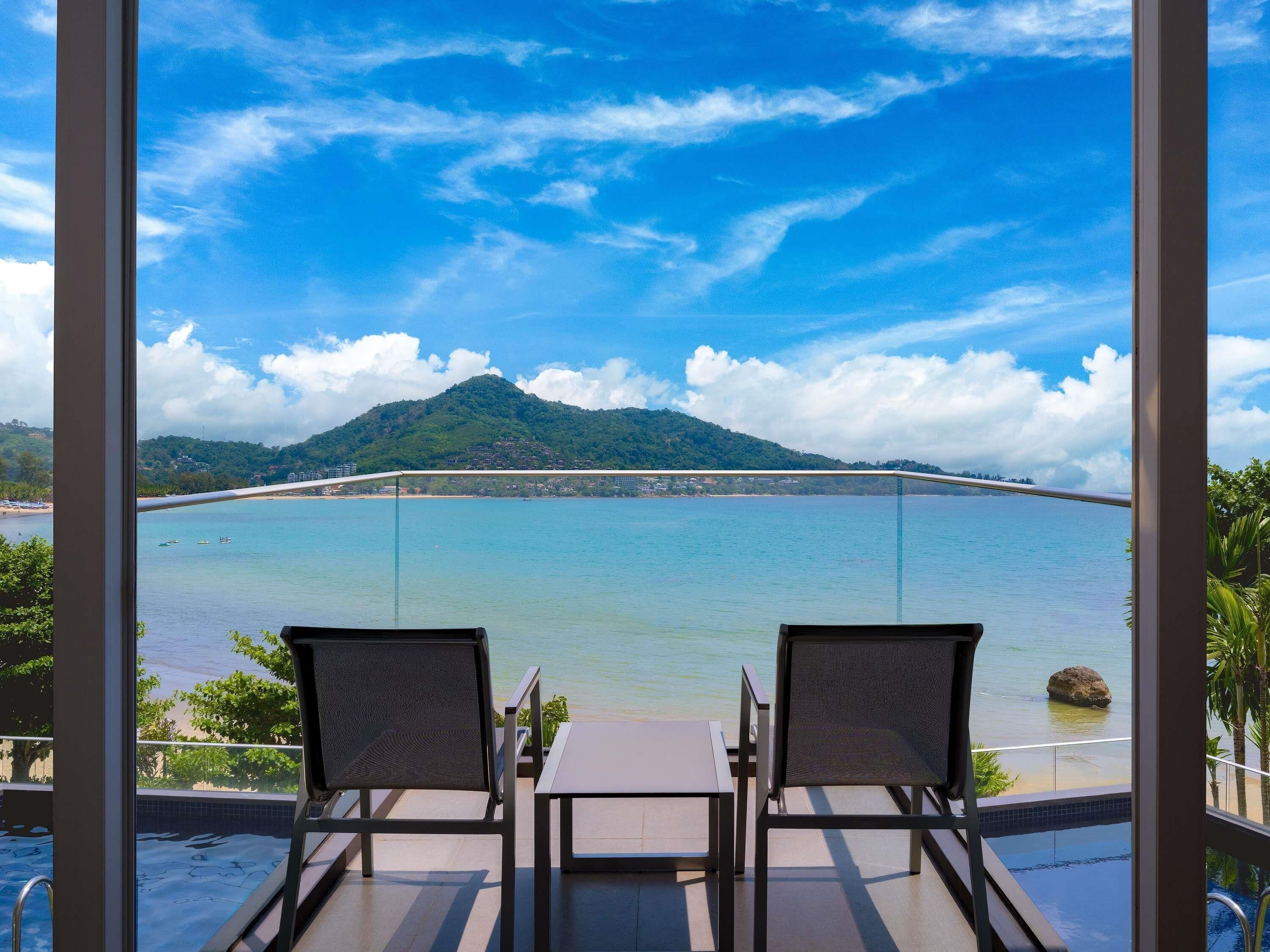 A balcony with two chairs and a table overlooking a body of water, a mountain, and a bridge.