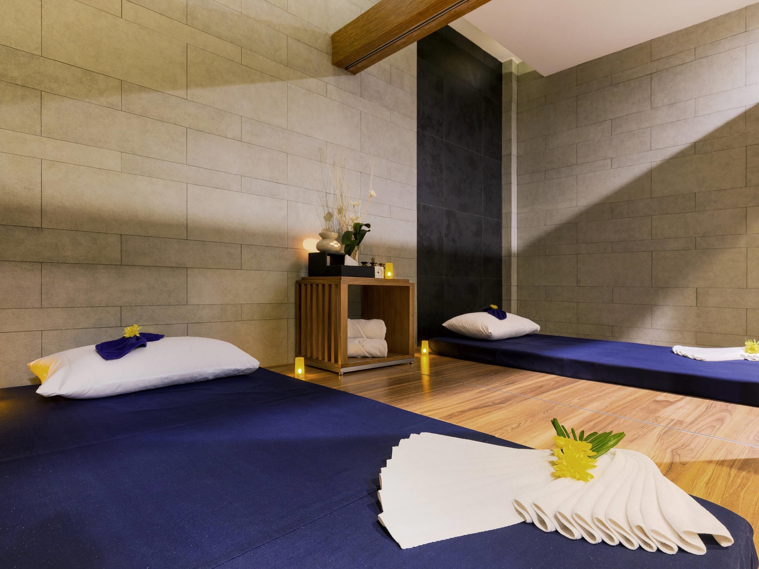 A spa room with two beds, a wooden cabinet with a vase of flowers, and a dark wall.