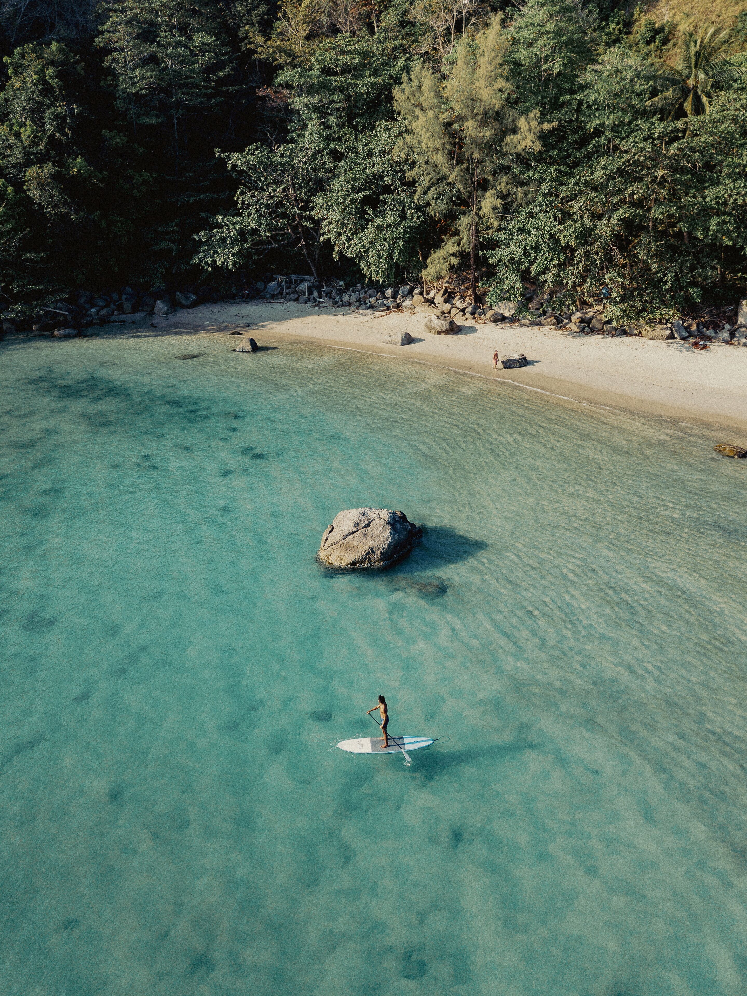 Un mar turquesa transparente con una persona haciendo paddleboard cerca de una roca grande.
