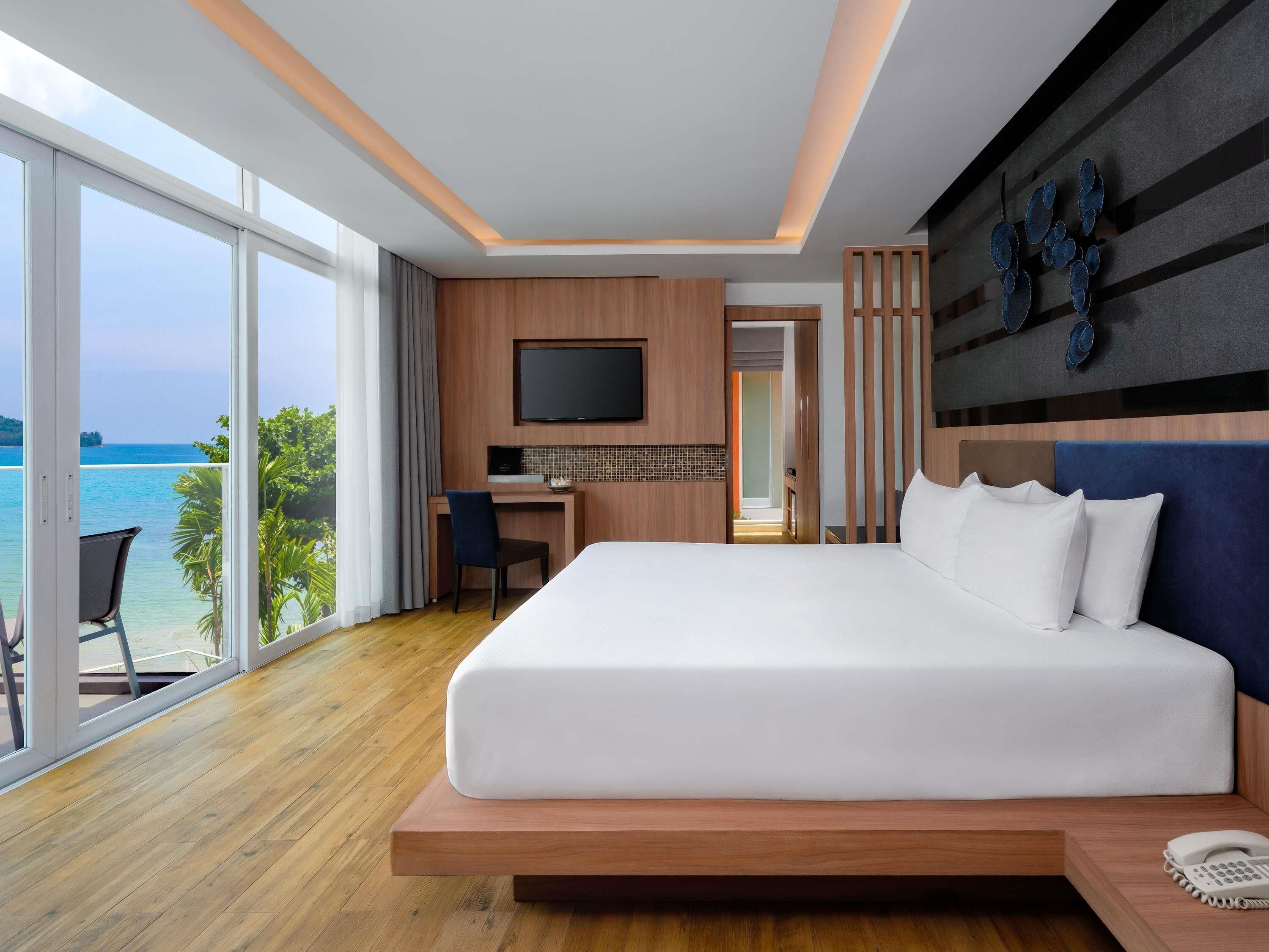 A modern hotel room with a large bed, a desk, a TV, and a view of the sea.