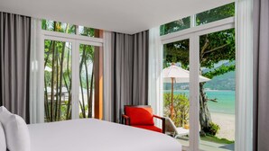 Habitación de hotel moderna con una cama grande, una silla roja y vistas a una playa y montañas a través de amplios ventanales.