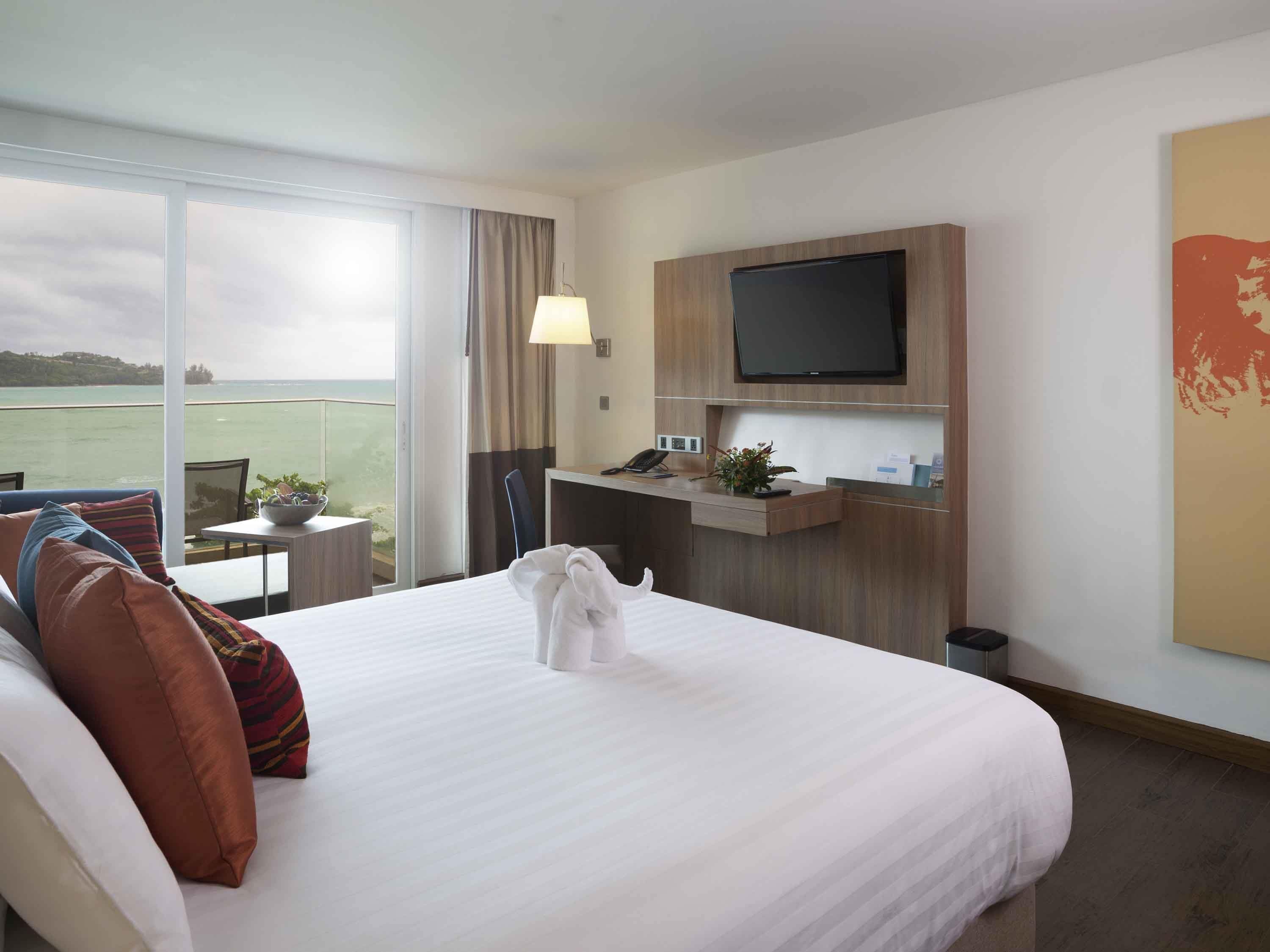 A hotel room with a bed, a desk, a TV, and a view of the sea.