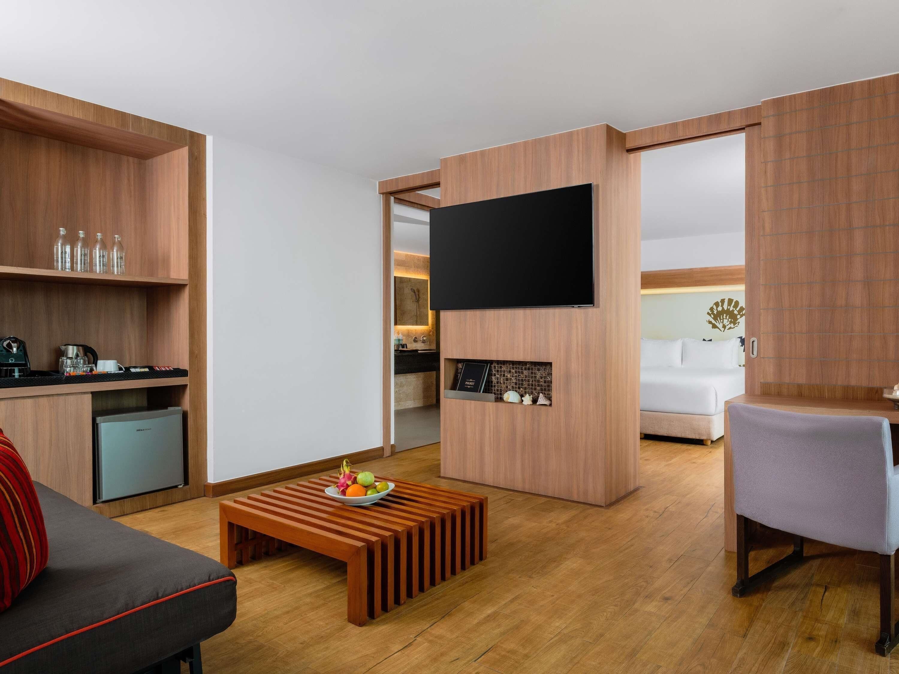A modern hotel room with a wooden interior, a flat-screen TV mounted on a wall, a kitchenette, a dining area with a table and chair, and a sofa.