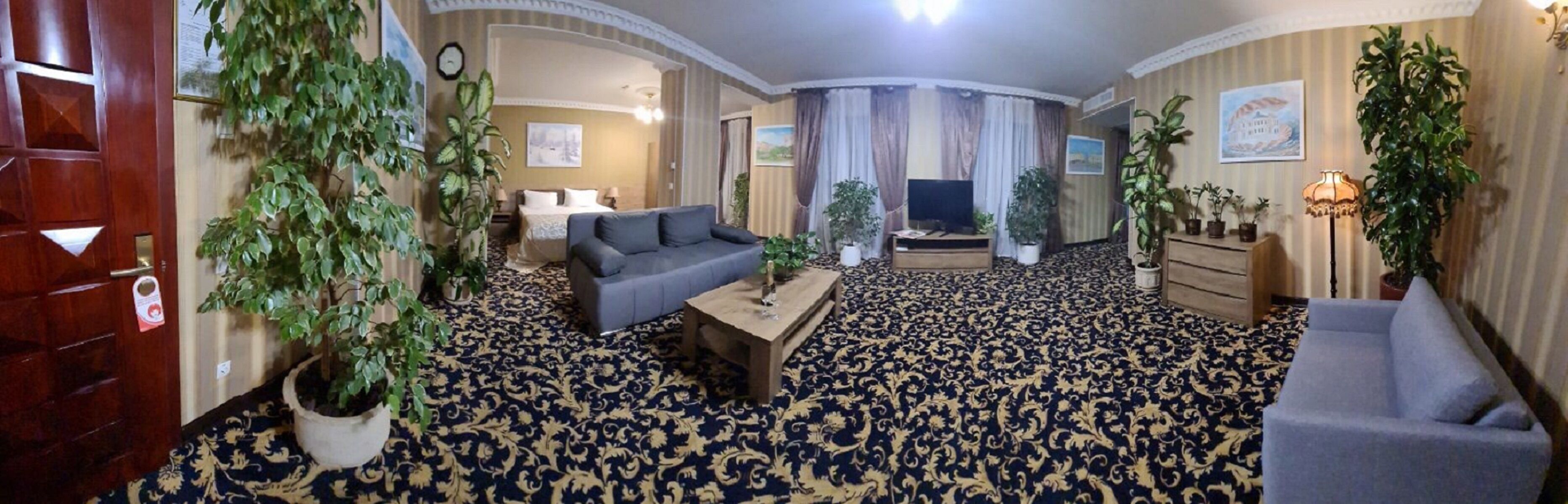 Una habitación de hotel con alfombra estampada, un sofá, una mesita de centro, un televisor y varias plantas en macetas.
