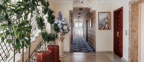 A hallway with potted plants, a chandelier, a blue carpet, and a painting on the wall.