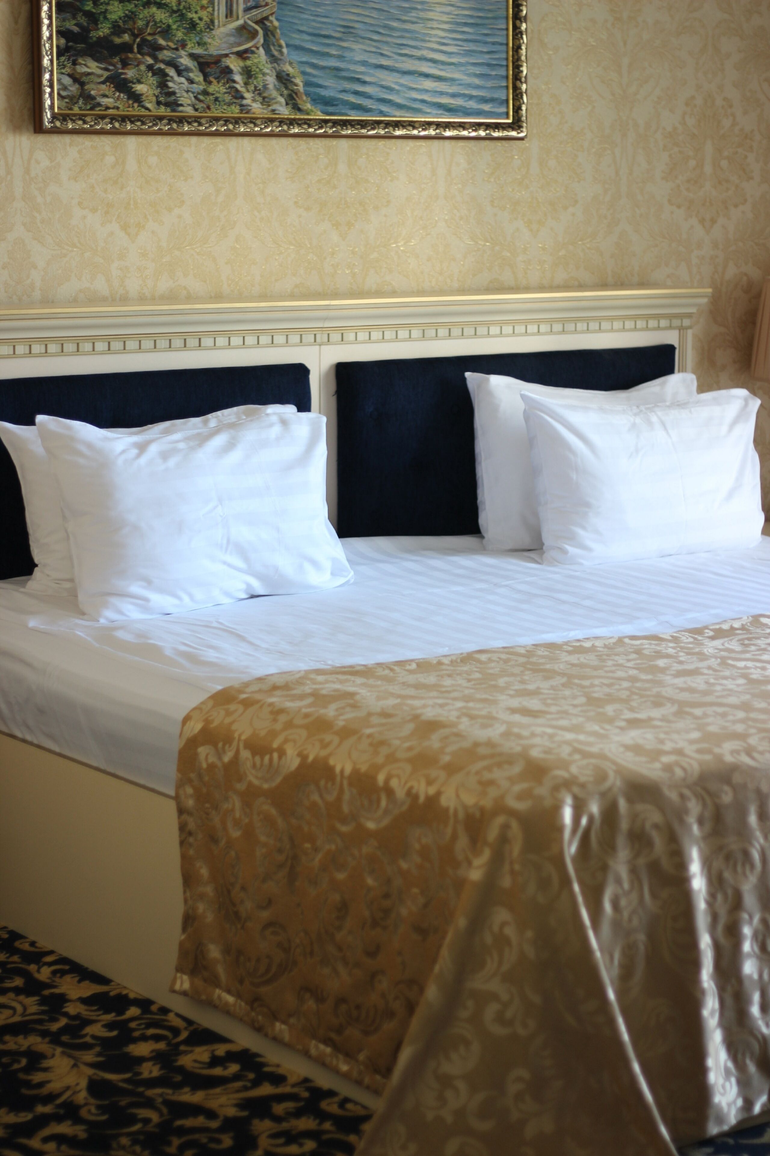 A neatly made bed with white and black pillows, a gold-patterned bedspread, and a framed painting above.