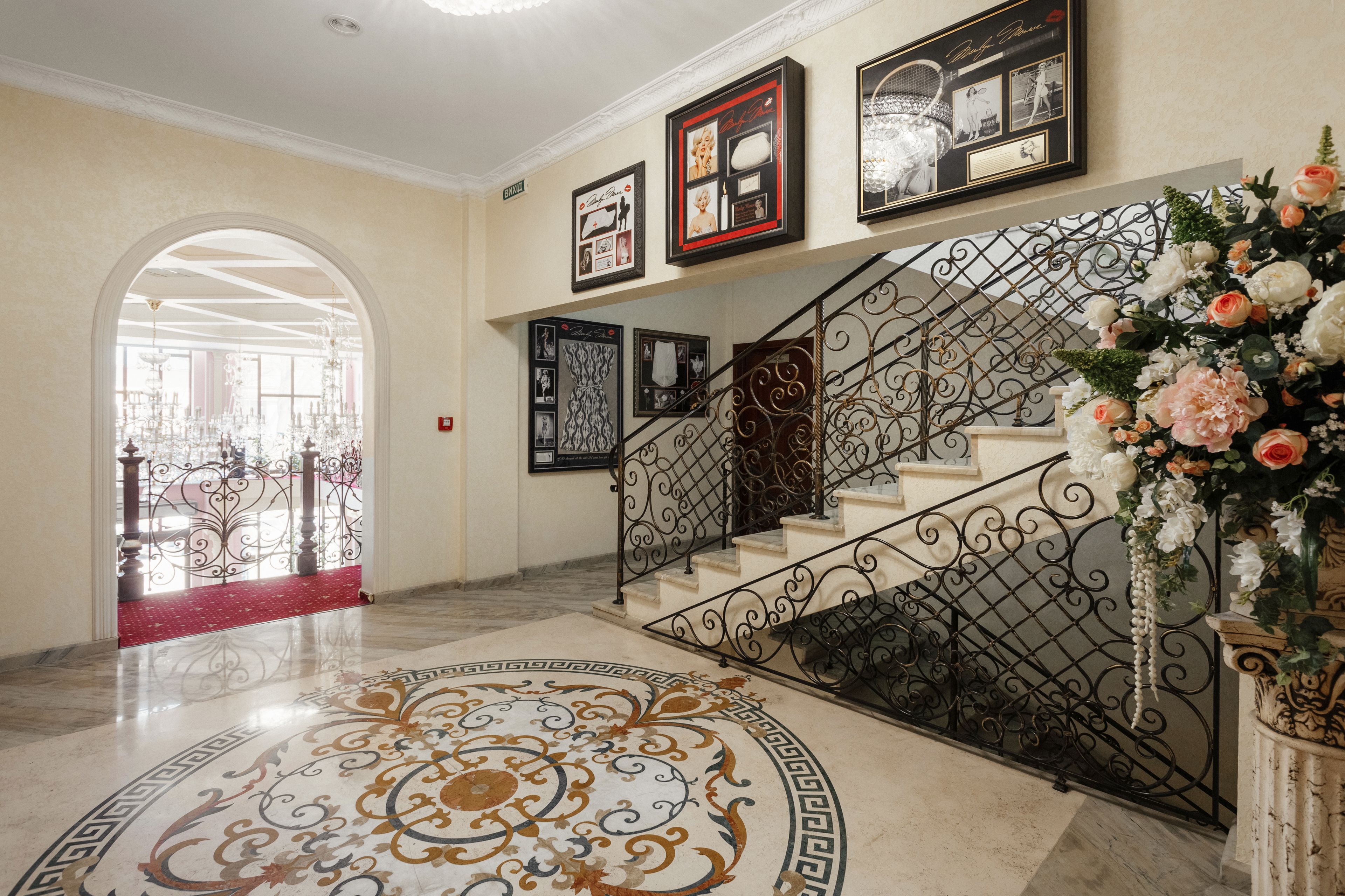 Una gran escalera con barandales de metal ornamentados, piso de mármol con diseños intrincados y un arreglo floral a la derecha.
