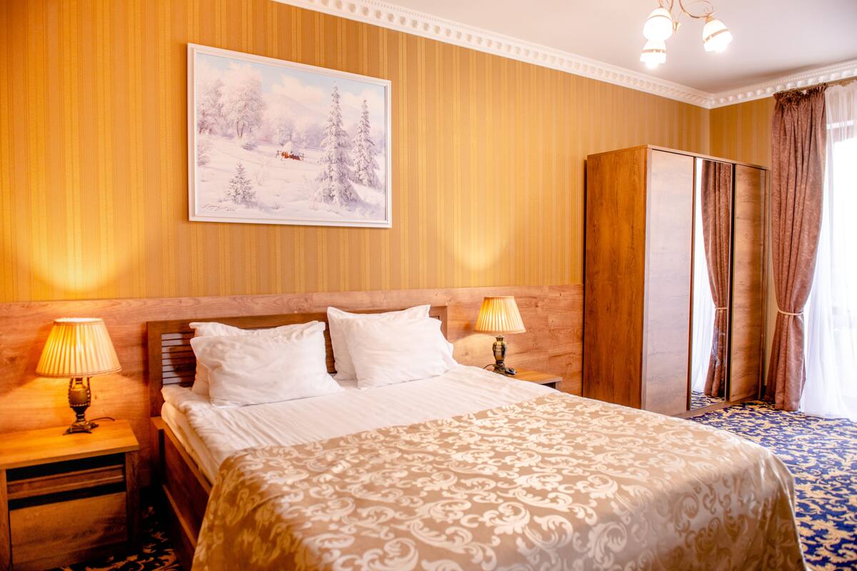 Hotellihuone, jossa on suuri sänky, kaksi yöpöytävalaisinta, puinen vaatekaappi ja seinällä oleva maalaus, jossa on lumipeitteinen maisema.