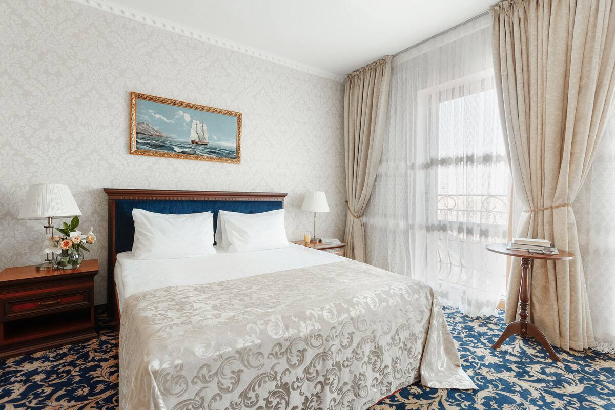 Hotellihuone, jossa on suuri sänky, kaksi yöpöytää, pieni sivupöytä, pieni pyöreä pöytä ja ikkuna, jossa on verhot.