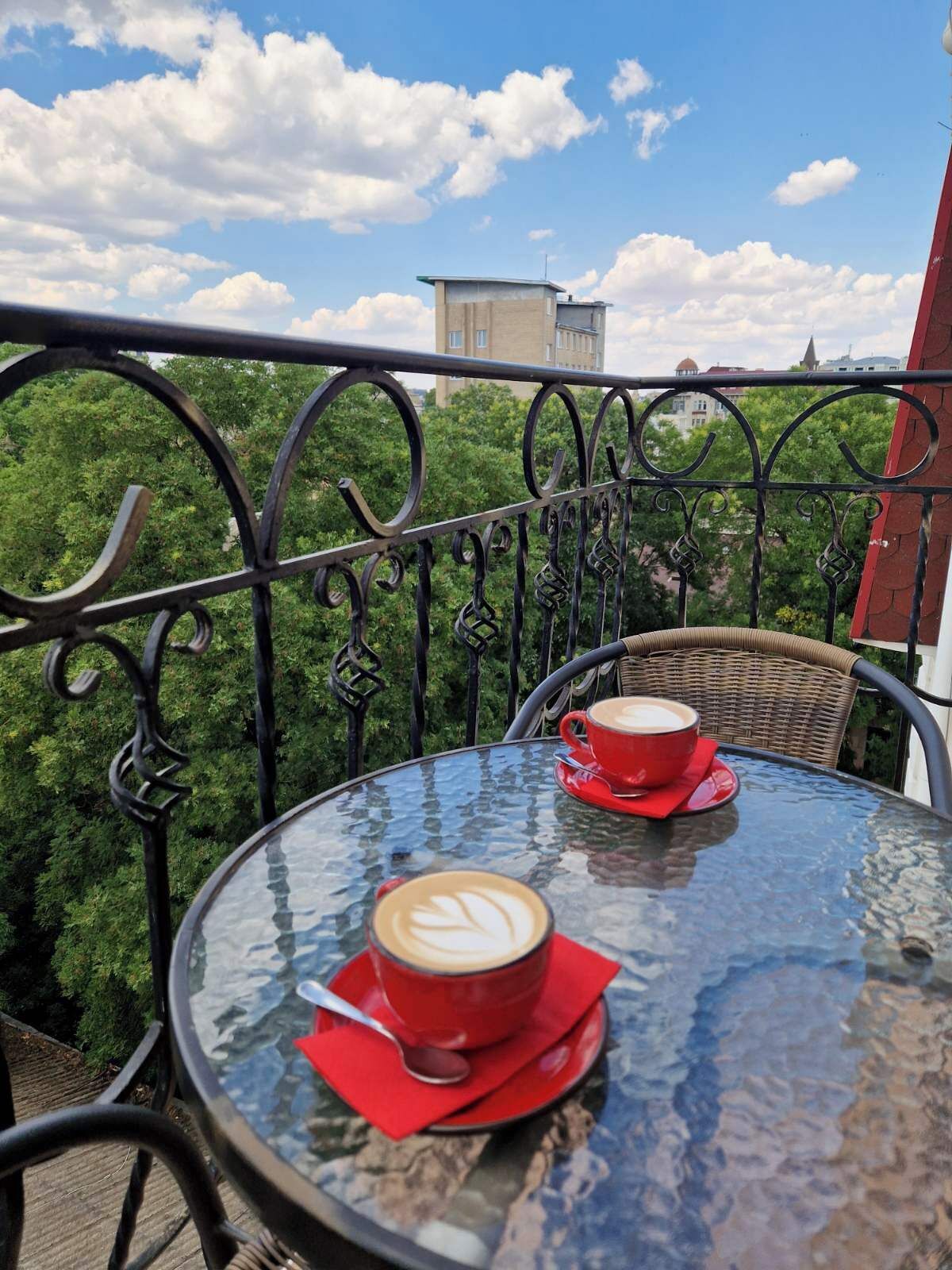 A round glass table with two red cups of coffee, a spoon, and a red napkin on a balcony with metal railings and greenery in the background.