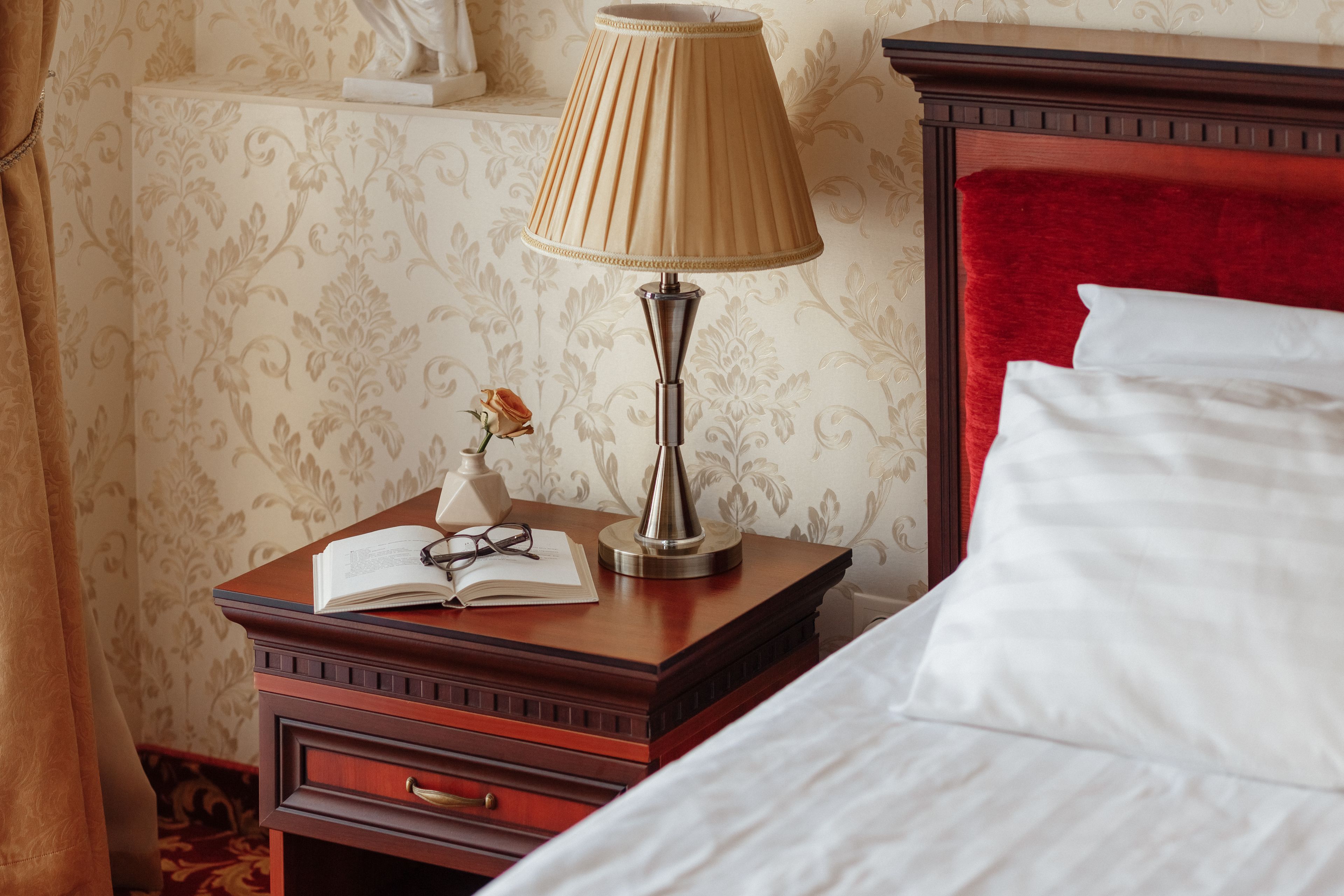 Una cama bien hecha con sábanas blancas, una mesita de noche de madera con una lámpara, un libro abierto y un par de gafas.