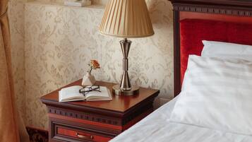 Una cama bien hecha con sábanas blancas, una mesita de noche de madera con una lámpara, un libro abierto y un par de gafas.