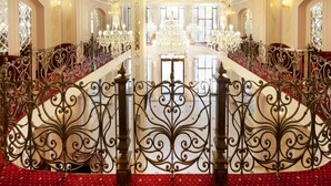 Un amplio hall de hotel con barandillas de hierro ornamentadas, una escalera central y un candelabro.
