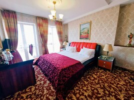 Habitación de hotel con una cama grande, un escritorio y una silla. Se ve una ventana con cortinas, un cuadro en la pared y una lámpara sobre el escritorio.
