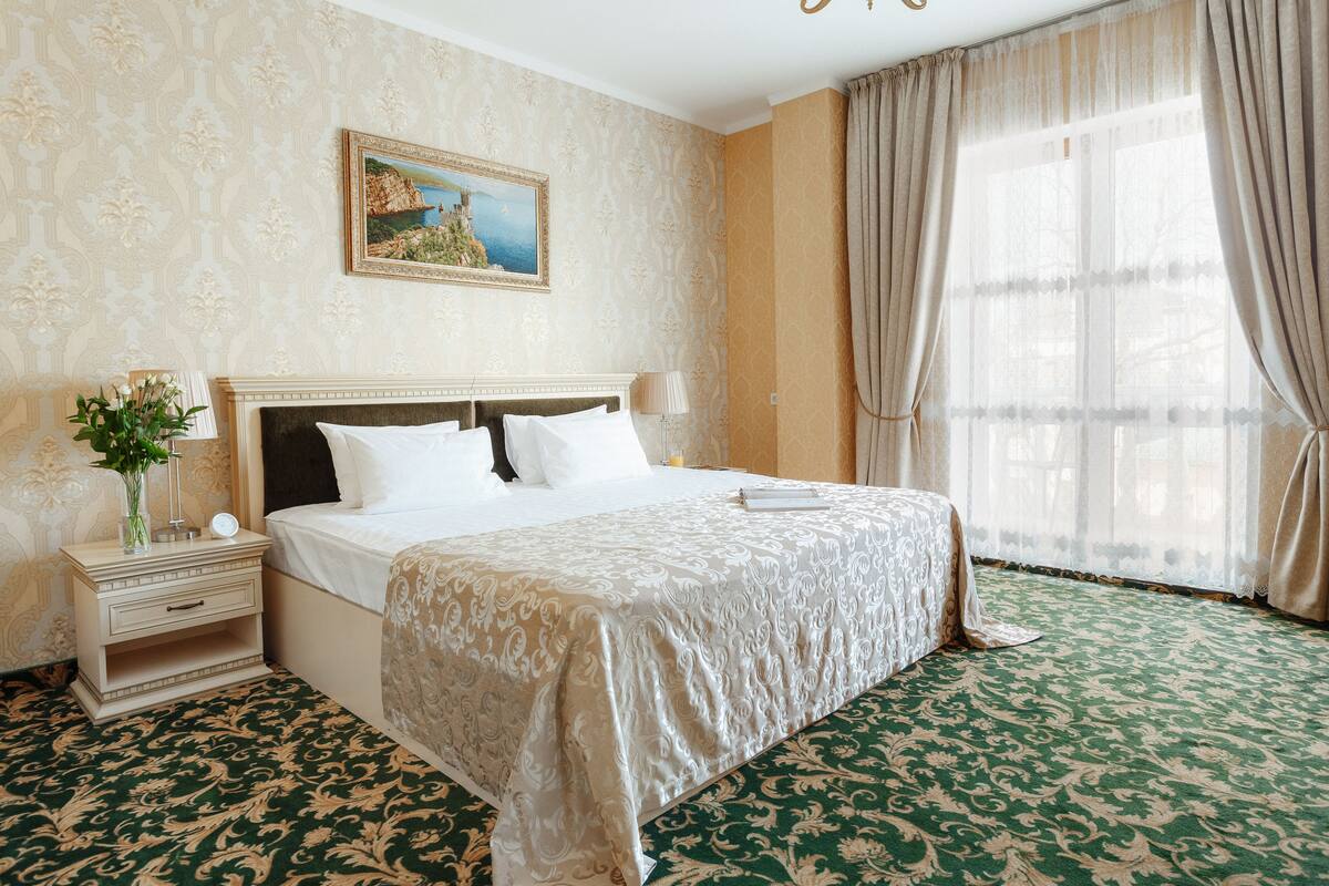 Hotellihuone, jossa on suuri sänky, yöpöytä lampun kanssa, taulu seinällä ja ikkuna, jossa on verhot.