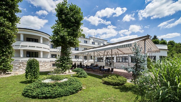 Un hotel con un diseño arquitectónico moderno, que cuenta con una zona de estar exterior cubierta con mesas y sillas, rodeada de jardines y árboles bien cuidados.