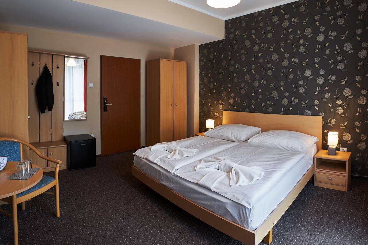 A hotel room with a single bed, a desk with a chair, a wardrobe, a small bedside table with a lamp, and a trash bin.