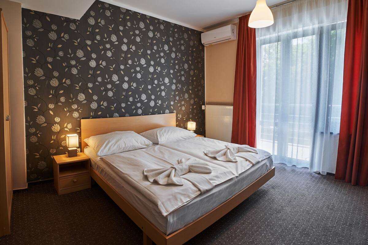 A hotel room with a large bed, a nightstand with a lamp, floral wallpaper, and a window with curtains.