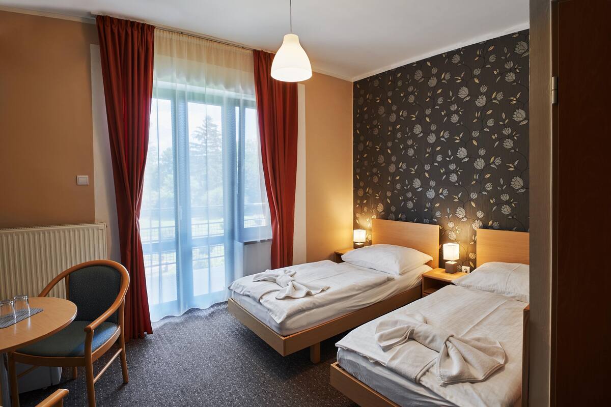 A hotel room with two single beds, a wooden chair, a table with glasses, a window with curtains, and a wall with a floral pattern.