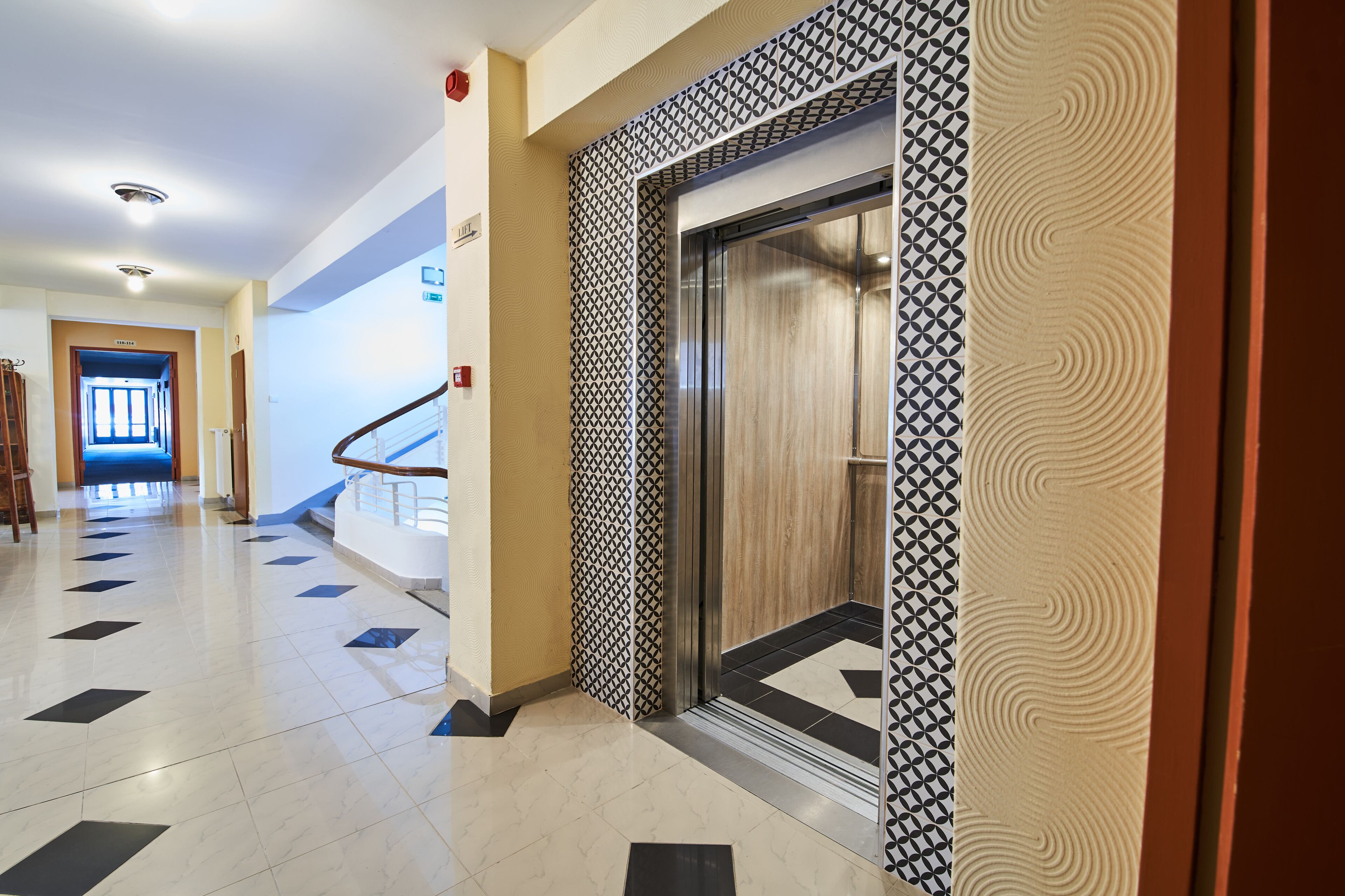 Un couloir d’hôtel moderne, avec un mur décoré de motifs, un ascenseur et un escalier.