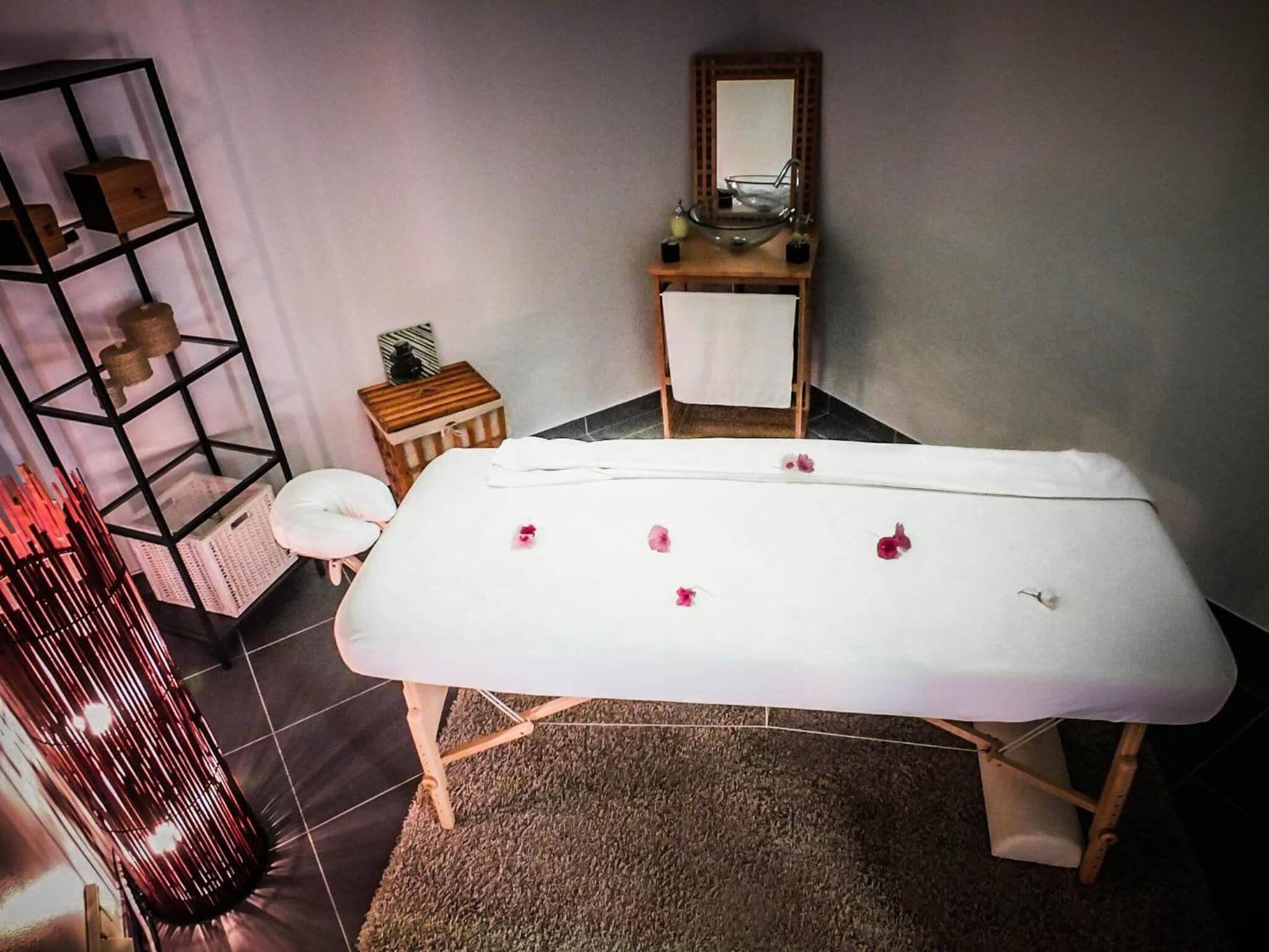 Una habitación con camilla de masaje, un mueble de madera pequeño, un espejo y una estantería con objetos decorativos.