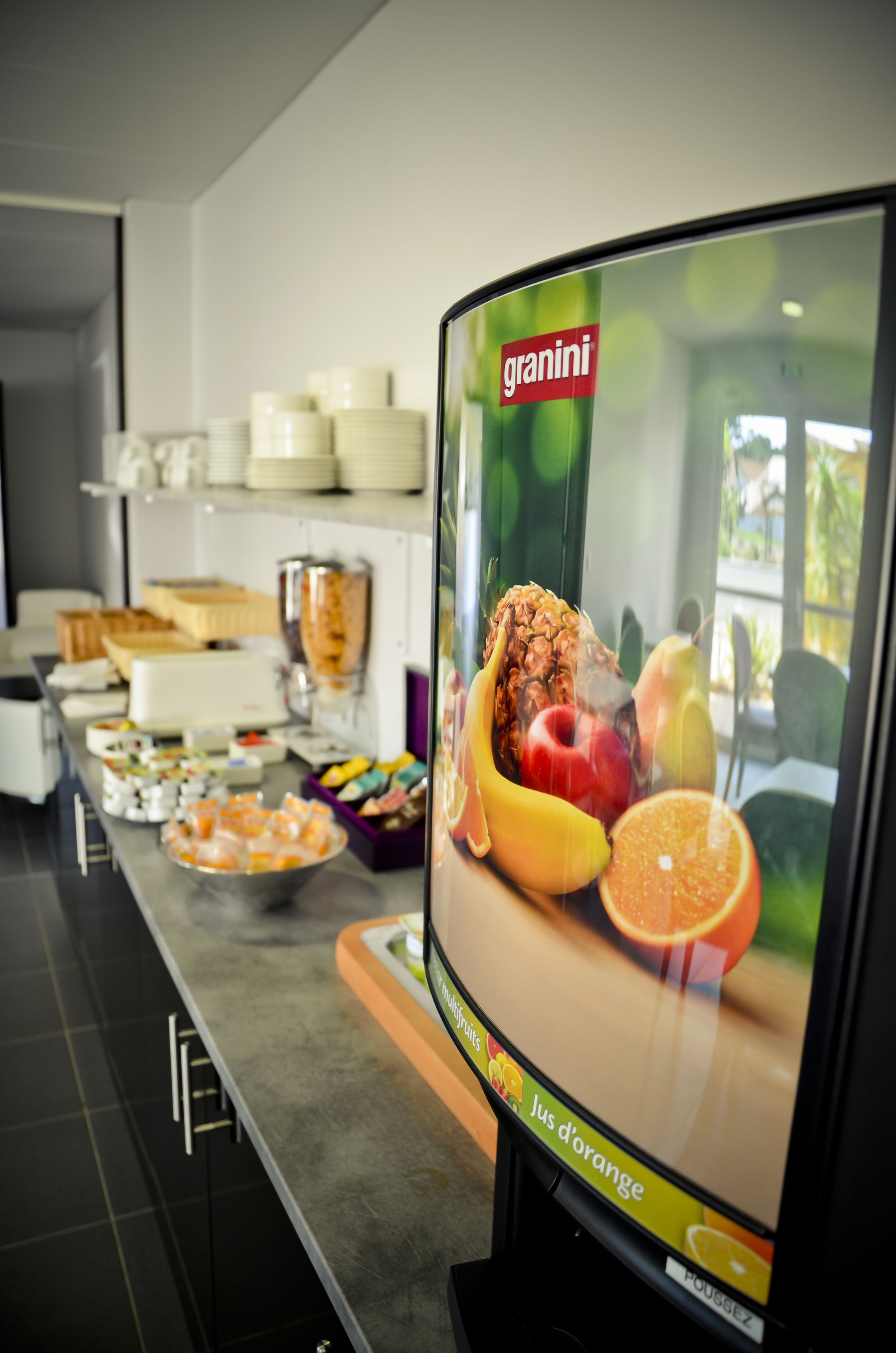 A buffet area with a television displaying a fruit advertisement, a variety of food items, and a stack of plates on a shelf.