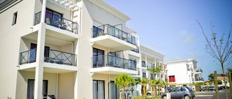 A multi-story residential building with balconies, a parking area, and small trees.