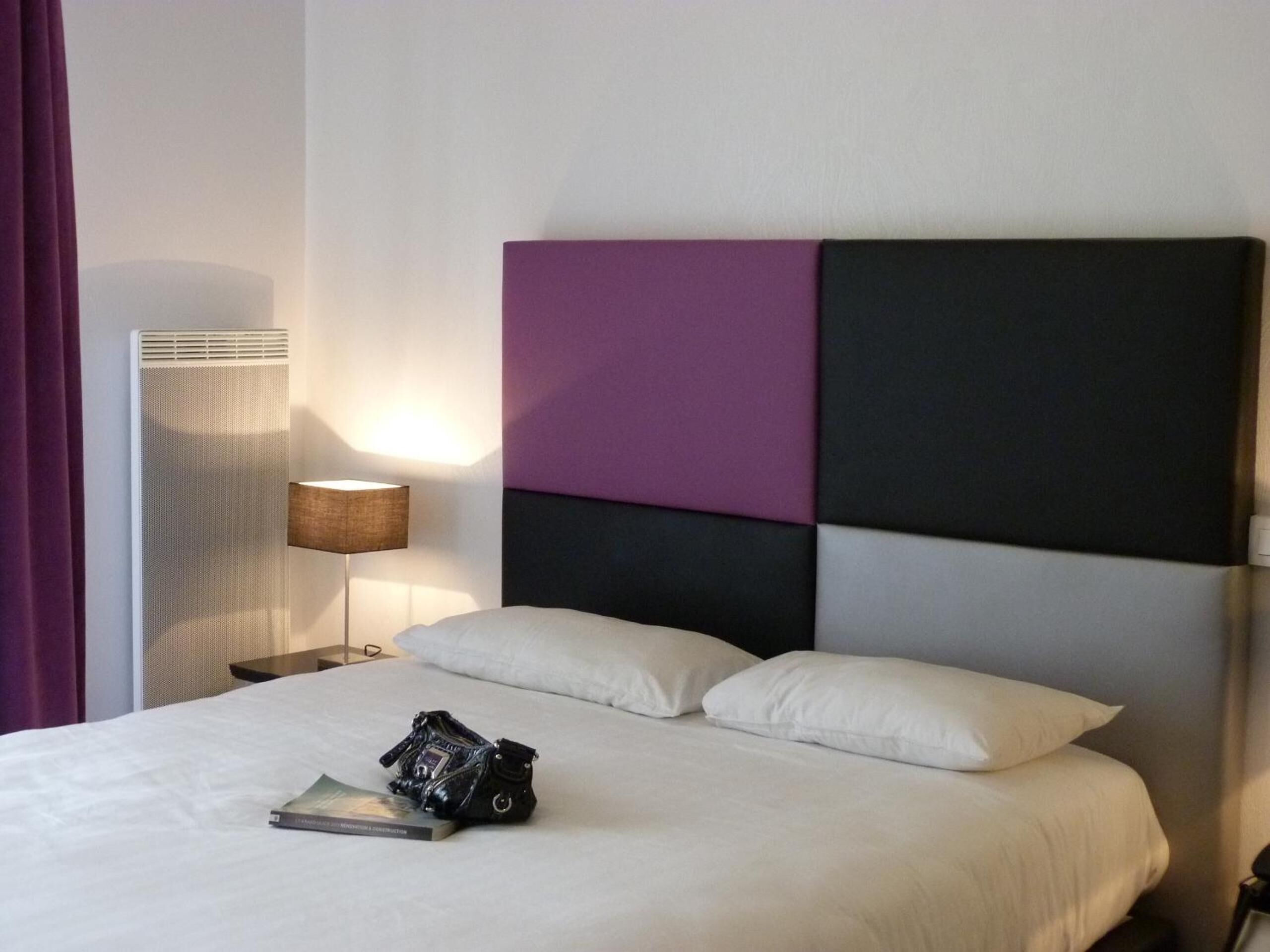 A hotel room with a bed, a nightstand with a lamp, and a wall unit with a purple and black color scheme.