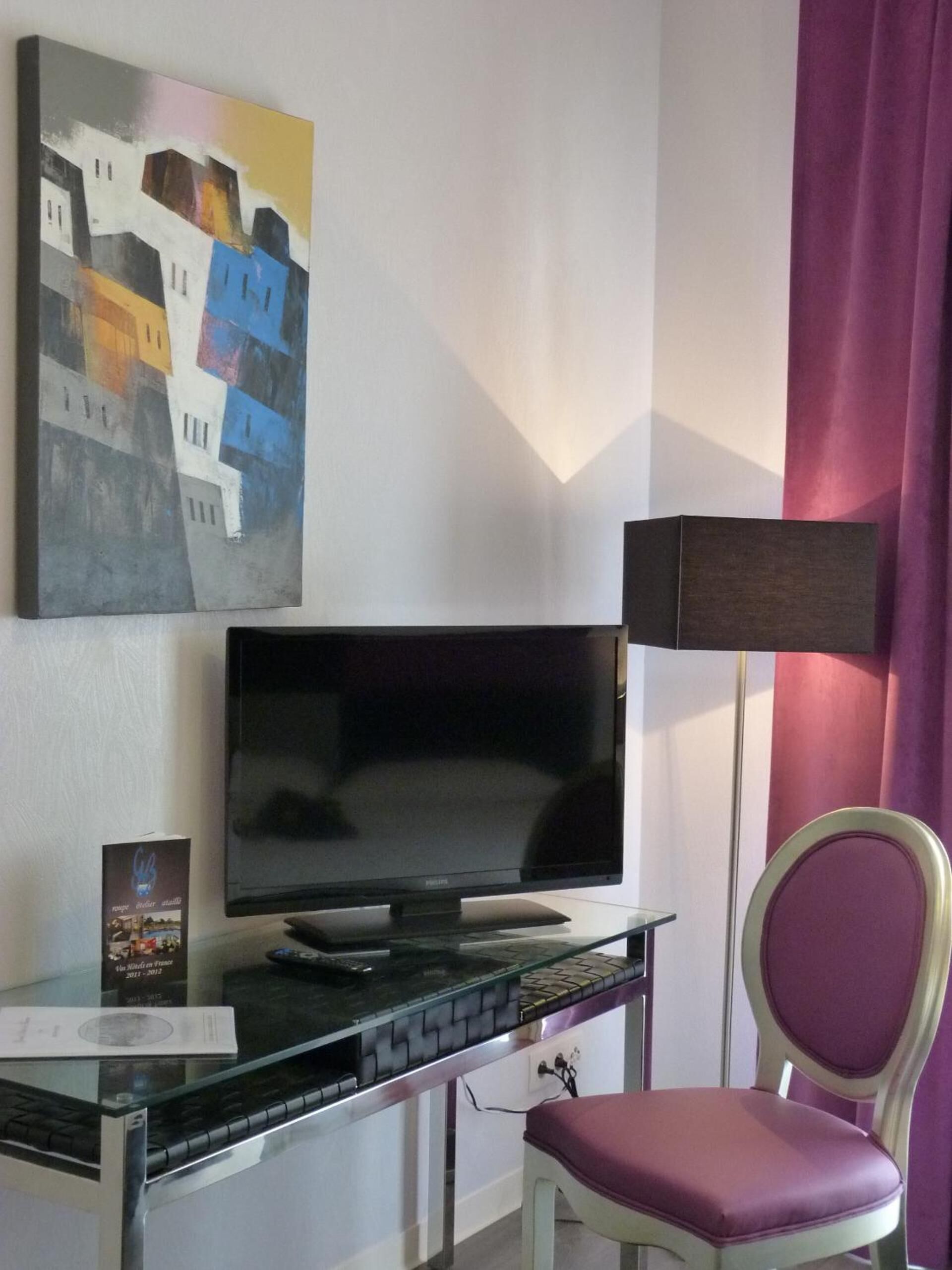 Et hotelværelse med et fladskærms-tv på et glastop, en stol med lilla pude, et billede på væggen og en lampe.