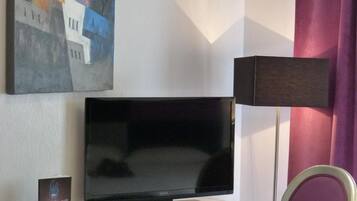Ein Hotelzimmer mit einem Flachbildfernseher auf einer Glastisch, ein Sessel mit violettem Kissen, einem Bild an der Wand und einer Lampe.