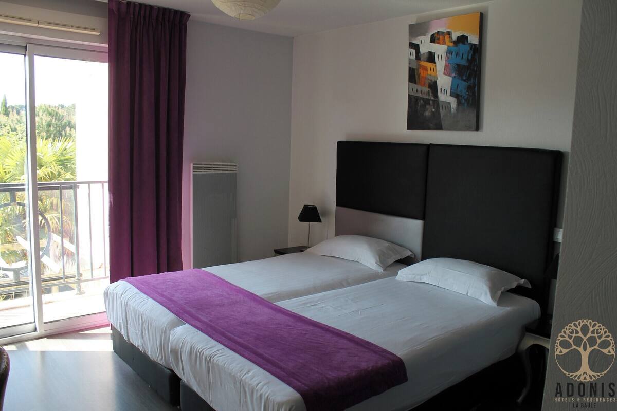 Quarto de hotel com cama grande, mesa de cabeceira, abajur e vista para o exterior.