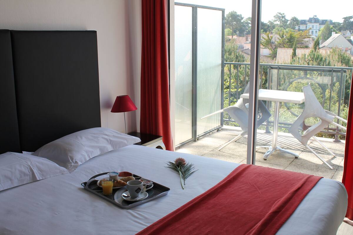 Quarto de hotel com cama, bandeja de café da manhã, cortina vermelha e varanda com cadeiras brancas.