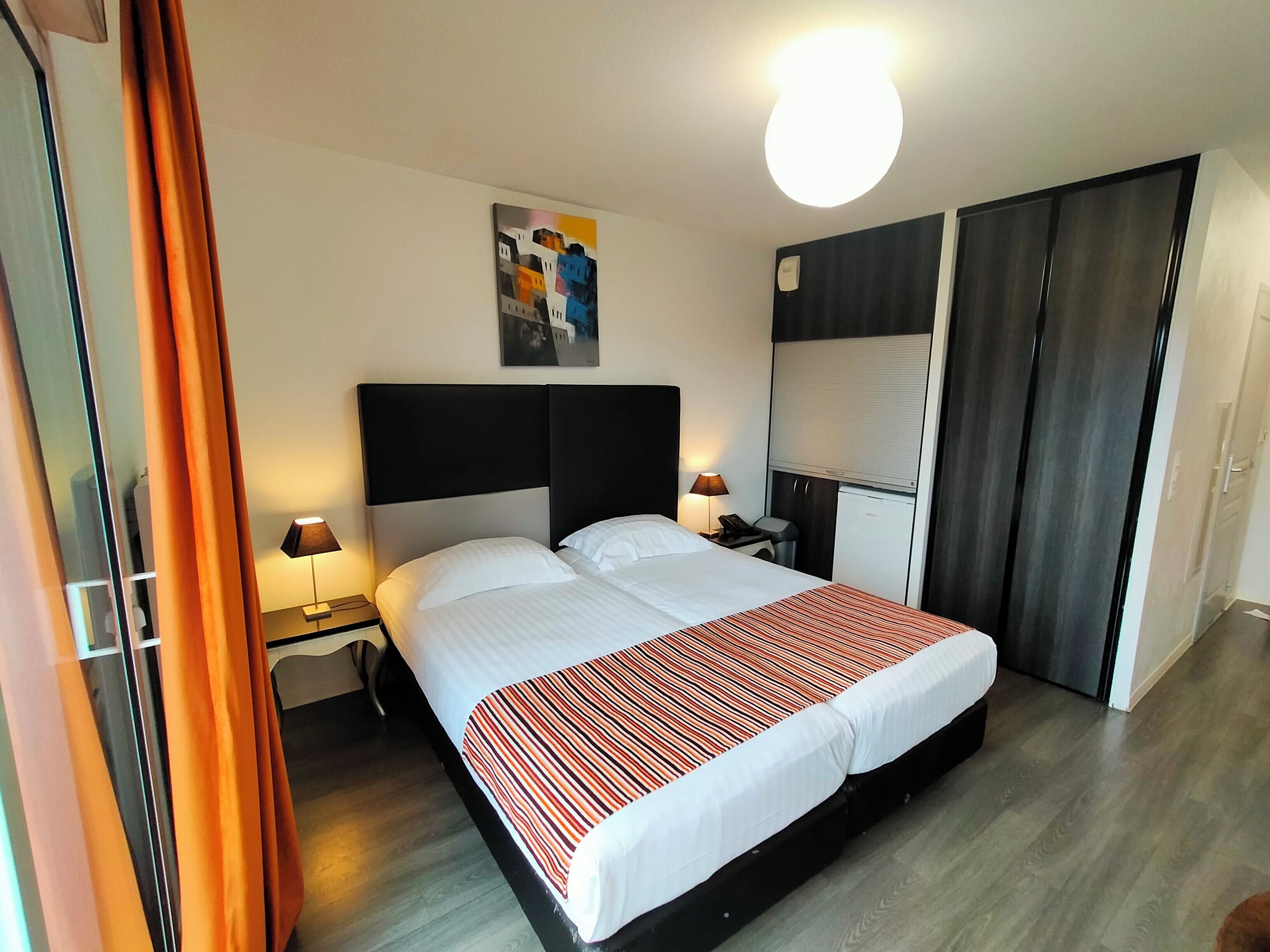Ett hotellrum med en säng, sänglampor, en tavla på väggen, en garderob och ett skrivbord med en lampa.