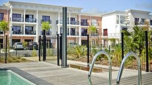 A swimming pool with a wooden deck, surrounded by a fence and a multi-story building with balconies. - Adonis La Baule (La Baule-Escoublac)