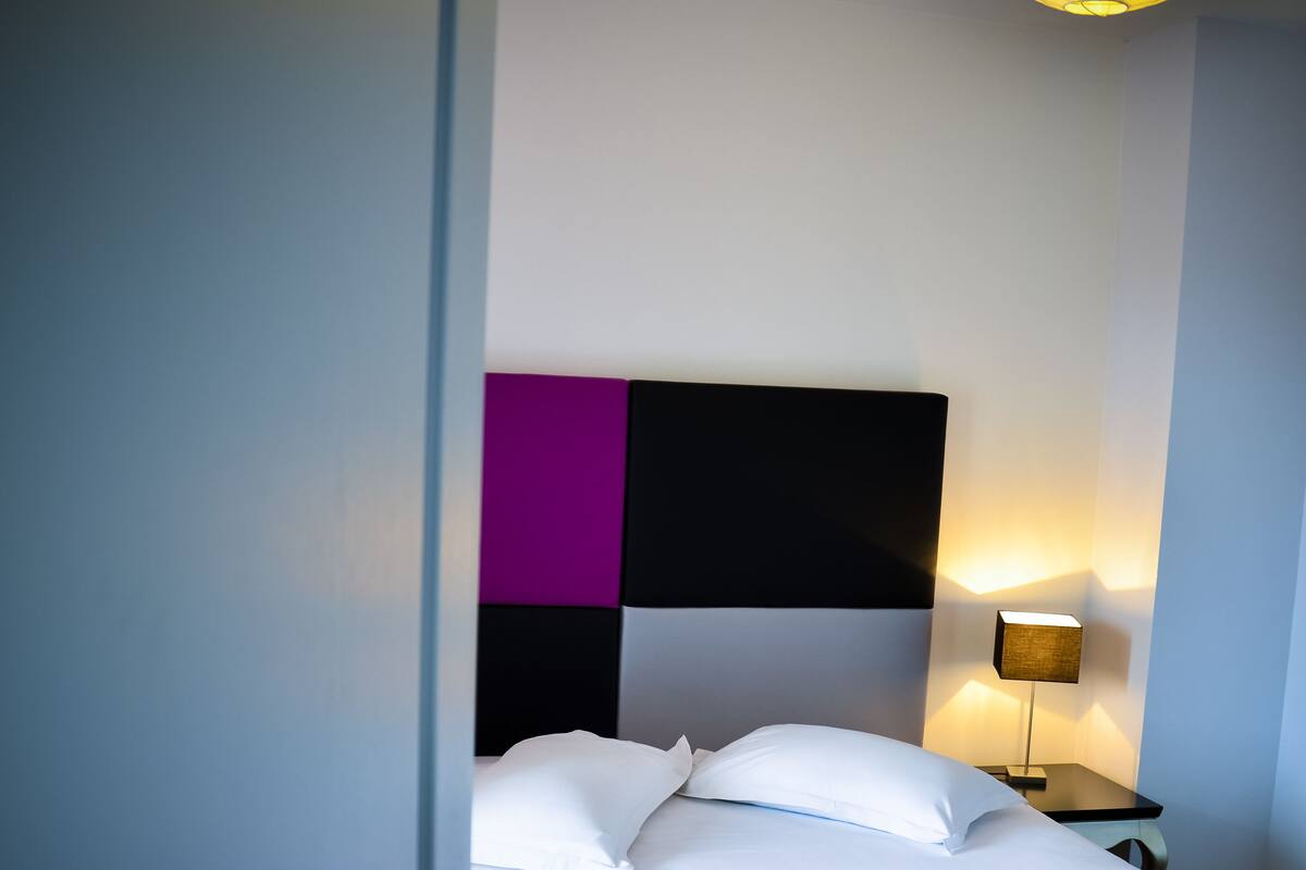 Cama bem arrumada com roupa de cama branca e roxa, cabeceira moderna e luminária de cabeceira.