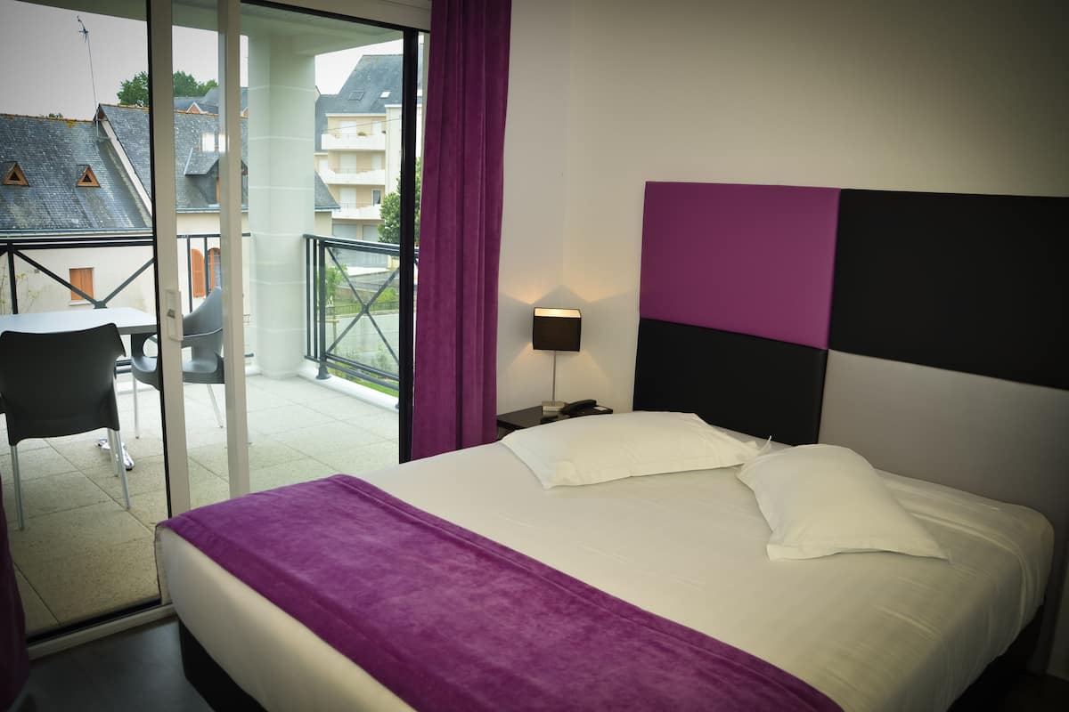 Quarto de hotel com cama, mesa de cabeceira, luminária, cadeira e varanda com vista para prédios.