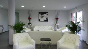 Um lobby moderno com um sofá branco, duas poltronas, uma mesa pequena e vasos decorativos com flores vermelhas.