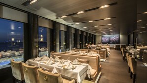 Um restaurante com várias mesas preparadas para refeições, amplas janelas com vista para a cidade e um design de interiores moderno.