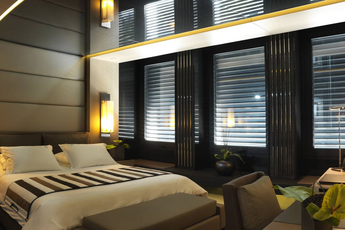 A modern hotel room with a large bed, a desk, and a chair. There are two windows with blinds, a nightstand, and a potted plant.
